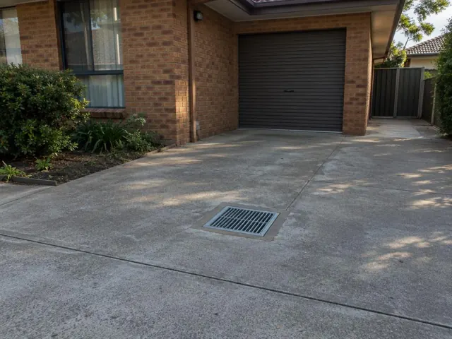 Concrete driveway with a square metal drain grate beside a brick house and garage door in Bonnyrigg